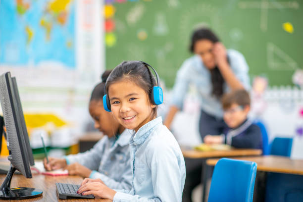 A small group of elementary children huddle together around a tablet as they watch a lesson electronically. They are each dressed casually and are focused on the screen.